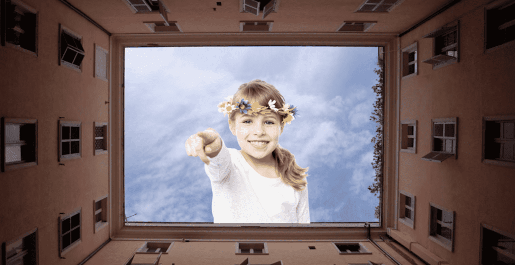 Little girl in the sky wearing long sleeve white shirt and flower crown points to camera with buildings bordering the frame