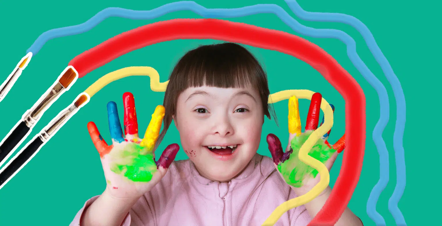 little girl smiles at camera with paint on her hands and fingers and paintbrushes in the background
