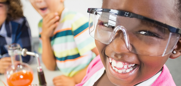 Little girl wearing transparent safety glasses smiles at the camera. A beaker with orange liquid is seen in the background and two other children post in the background (their faces cropped out)