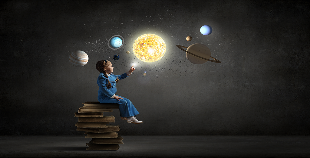 A young girl sits on a stack of books and points to the solar system lit up above and in front of her