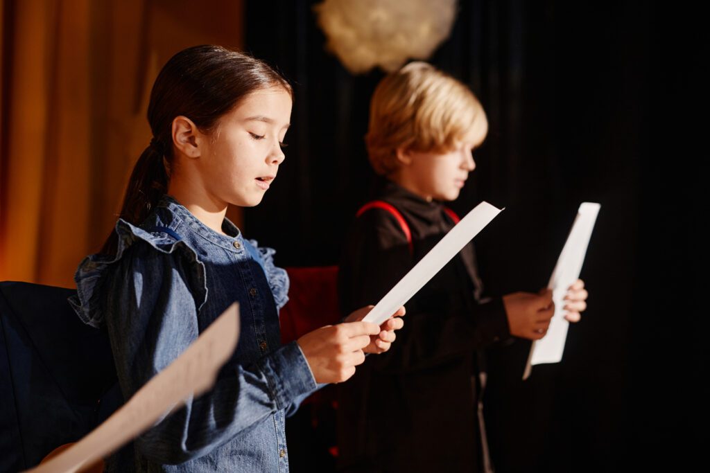 two children stand with papers in their hands on a stage