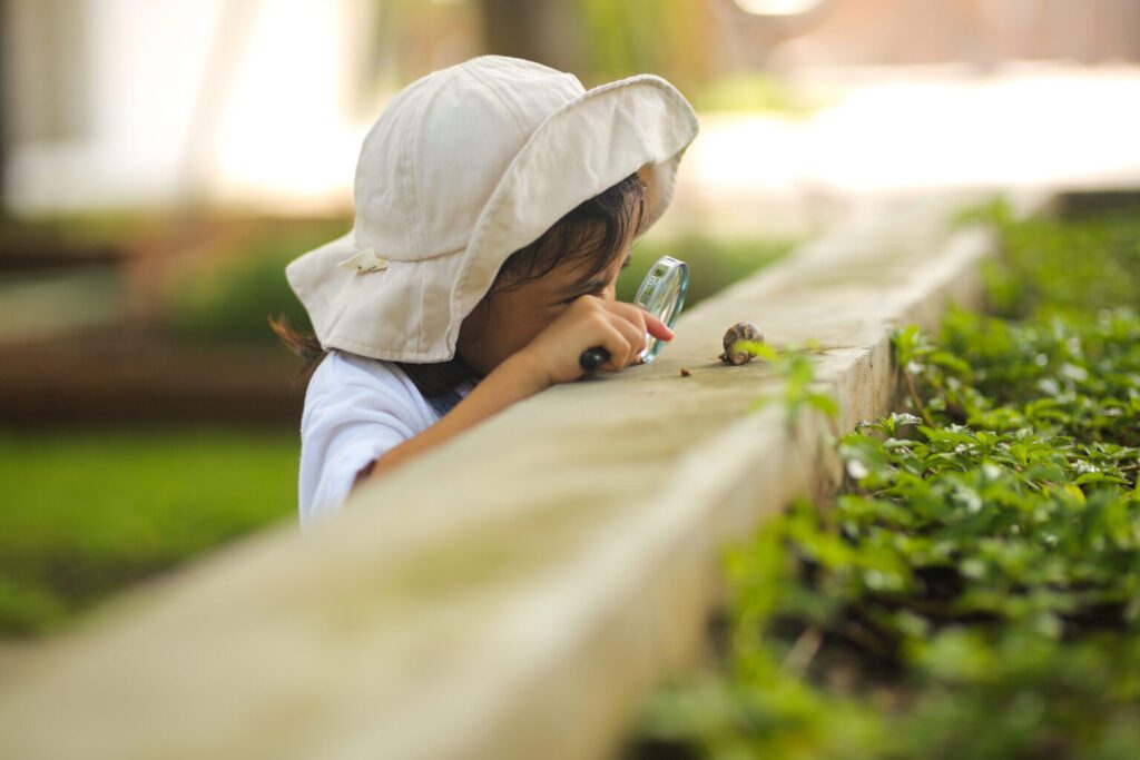 Little kid girl wearing a white hat and jeans jumpsuit and exploring nature with a magnifying glass.