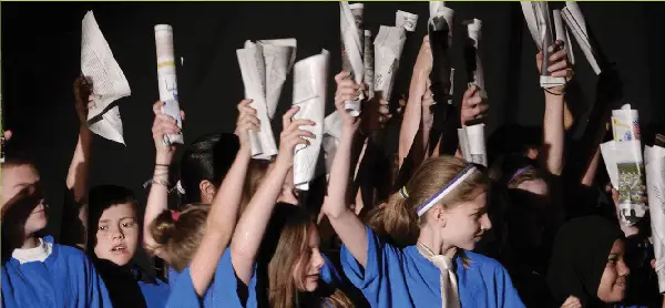 Kids in blue shirts hold up white newspapers