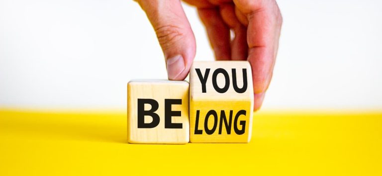 you belong in Black letters on a light brown blocks on a yellow table with white background. A hand grabs one of the blocks